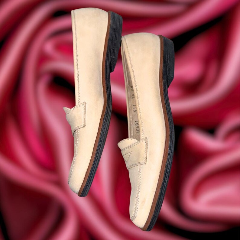 Puede incluir: Un par de mocasines de cuero beige con un dise&ntilde;o cl&aacute;sico. Los zapatos tienen una suela marr&oacute;n oscuro y una correa de sill&iacute;n de color claro en la parte superior. Los zapatos se muestran sobre un fondo rosa borroso.
