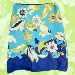 Puede incluir: Falda en forma de A con un vibrante estampado floral. La falda presenta un fondo azul claro con grandes diseños florales blancos, verdes y azul marino. El dobladillo es azul marino liso. La falda está colgada de una percha plateada.