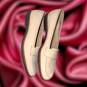 Puede incluir: Un par de mocasines de cuero beige con un dise&ntilde;o cl&aacute;sico. Los zapatos tienen una puntera redondeada, una correa de centavo y una suela marr&oacute;n oscuro. Las costuras son visibles y los zapatos parecen nuevos.