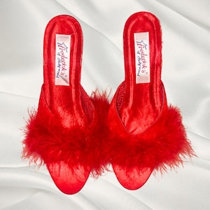 Puede incluir: Un par de zapatillas de satén rojo con adornos de plumas. Las zapatillas tienen una punta puntiaguda y un tacón pequeño. El nombre de la marca "Frederick's of Hollywood" está impreso en la plantilla.