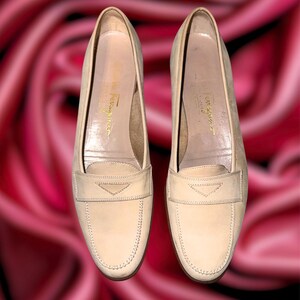 Puede incluir: Un par de mocasines color crema con tac&oacute;n bajo. Los zapatos tienen un dise&ntilde;o cl&aacute;sico con una correa decorativa en la parte superior y detalles de costura. El nombre de la marca es visible en la plantilla.