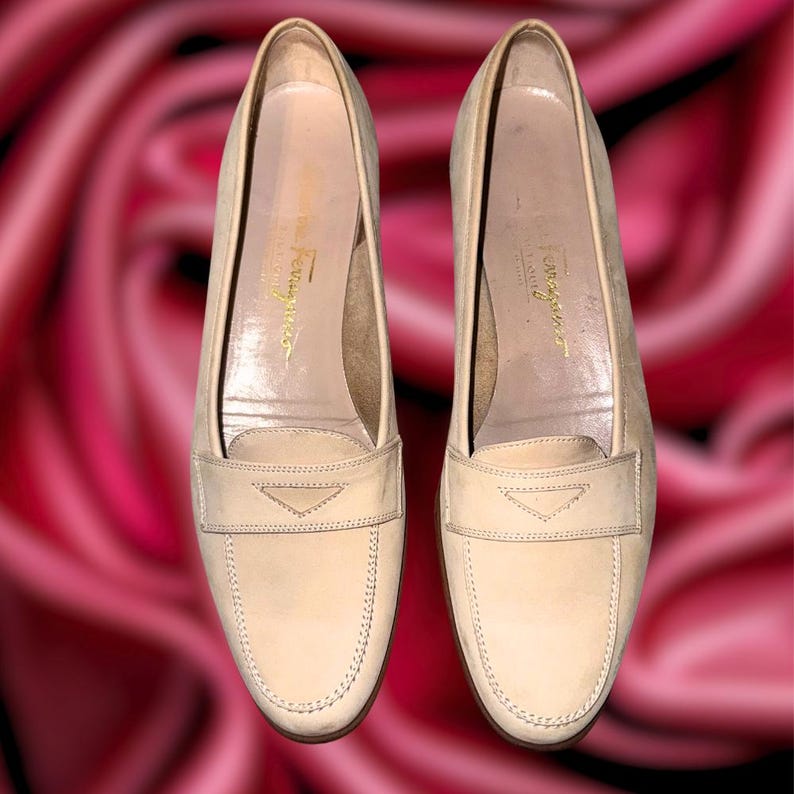 Puede incluir: Un par de mocasines color crema con tac&oacute;n bajo. Los zapatos tienen un dise&ntilde;o cl&aacute;sico con una correa decorativa en la parte superior y detalles de costura. El nombre de la marca es visible en la plantilla.