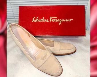 May include: A pair of beige leather loafers with a low heel, displayed with a red Salvatore Ferragamo box. The shoes have a classic design with a stitched vamp and a decorative strap across the top. The box features the brand's logo in gold.