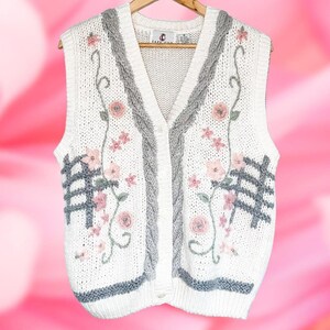Puede incluir: Un chaleco de punto crema con botones y bordados florales. El chaleco presenta flores rosas, detalles grises y un diseño de punto trenzado en los bordes. El chaleco es sin mangas y cuelga de una percha de madera.