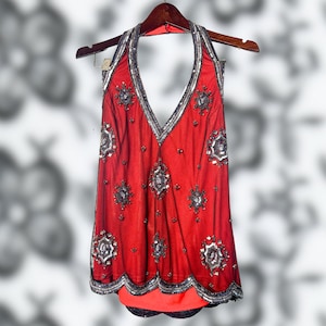 May include: A red halter top with silver and black sequin embellishments. The top features a V-neckline and is adorned with intricate sequin patterns. It hangs on a wooden hanger, showcasing its design and details. The garment's bottom edge has a scalloped design.