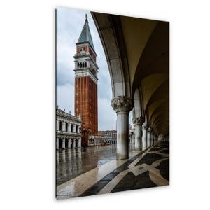 Peut inclure: Photographie de la Campanile de Saint-Marc à Venise, Italie, une haute tour de clocher en briques avec une flèche verte. L'image montre également une colonnade avec des plafonds voûtés et un sol à motifs, reflétant le pavé mouillé.