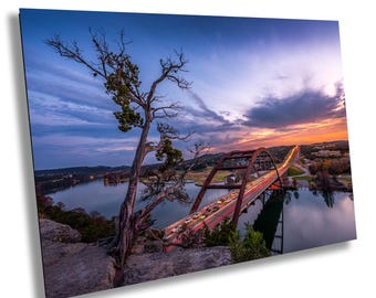 May include: A scenic view of a bridge over a lake at sunset. The bridge is lit up with car headlights, creating a trail of light. A tree in the foreground frames the scene.