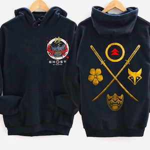 May include: Black hooded sweatshirt with a gold and red graphic of a samurai mask and the text "Ghost of Tsushima" on the front. The back of the sweatshirt features a gold graphic of two crossed swords, a flower, a fox, and a mask.