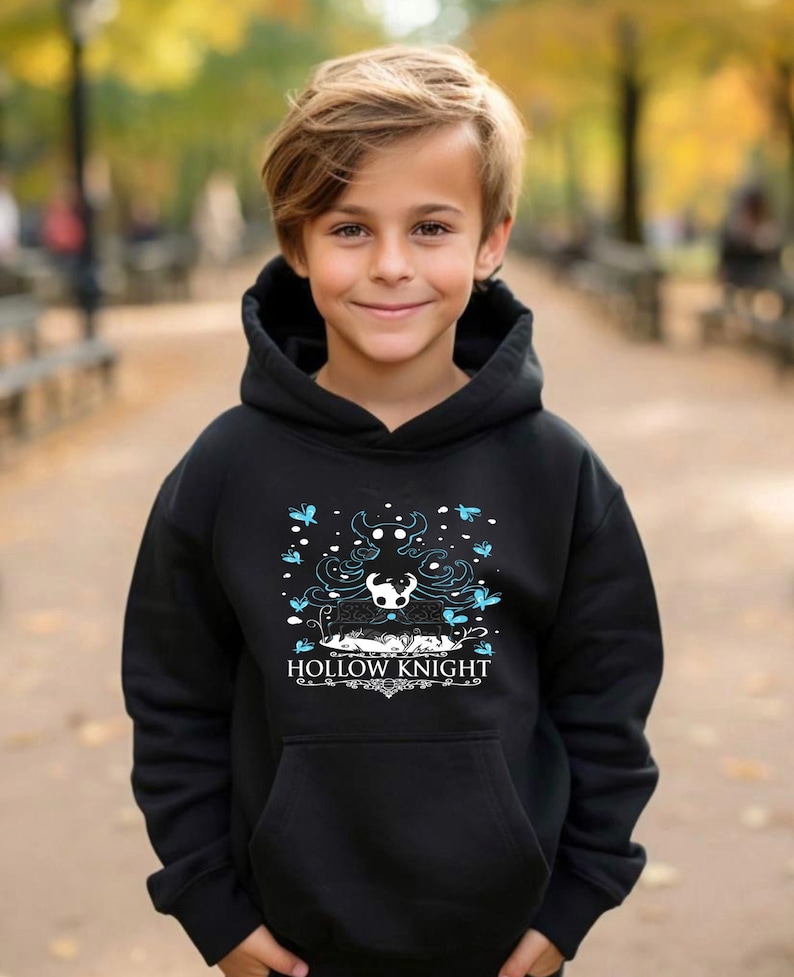 Pode incluir: Camisola de capuz preta com um gr&aacute;fico branco de um personagem do videojogo Hollow Knight. O gr&aacute;fico inclui o texto "Hollow Knight" e um desenho do personagem com uma coroa e asas.