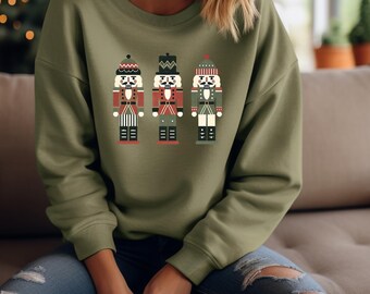 May include: An olive green sweatshirt featuring a graphic of three toy nutcrackers in red, black, and white. The nutcrackers have different hats and uniforms. The sweatshirt is a long-sleeved crew neck.