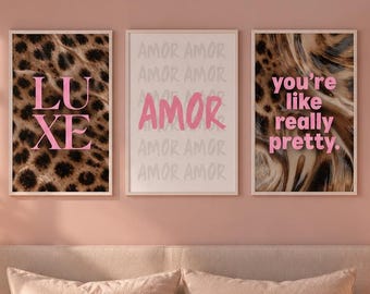 May include: Three rectangular wall art prints in white frames hanging above a bed. The left print features a leopard print background with the text LUXE. The center print has the word AMOR in pink. The right print features the text you're like really pretty.