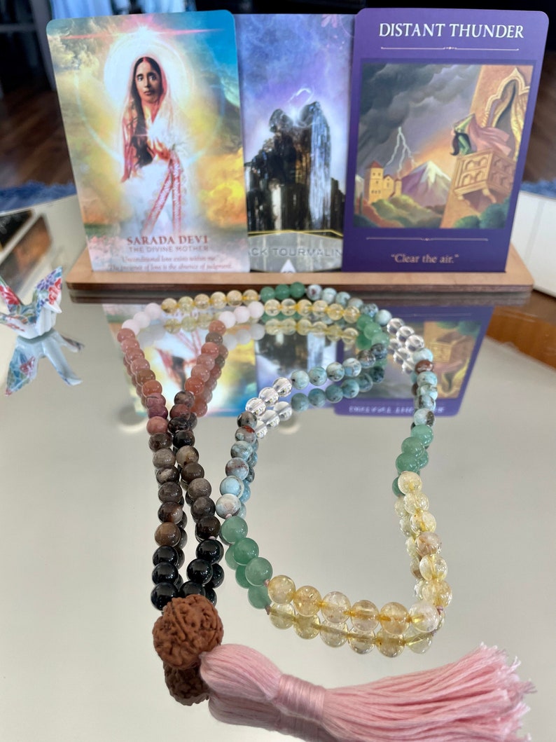 May include: A multi-colored beaded mala necklace with a pink tassel. The mala is made of various stones, including clear quartz, green aventurine, and brown jasper. The mala is laid out on a reflective surface with three tarot cards in the background. The cards are "Sarada Devi", "Black Tourmaline", and "Distant Thunder".