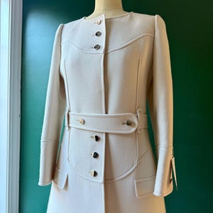 May include: A white long-sleeved coat with a belt and gold buttons. The coat has a fitted waist and a flared skirt.