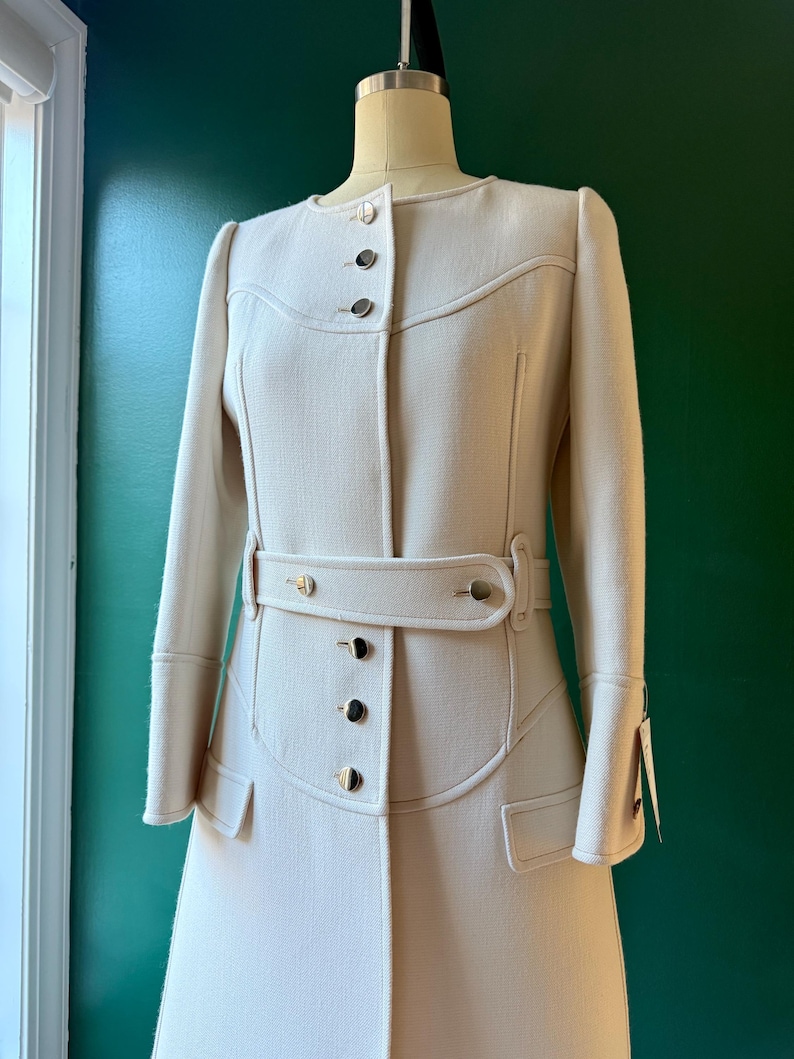 May include: A white long-sleeved coat with a belt and gold buttons. The coat has a fitted waist and a flared skirt.