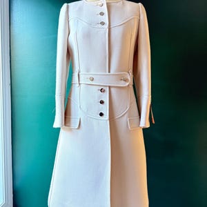 May include: A long, white coat with a belted waist and a button-up front. The coat has a slightly flared skirt and long sleeves.