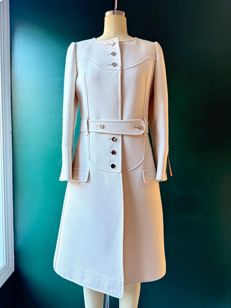 May include: A long, white coat with a belted waist and a button-up front. The coat has a slightly flared skirt and long sleeves.
