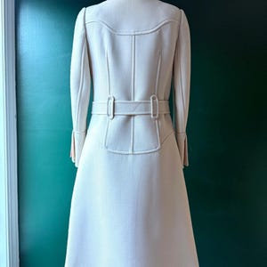 May include: A white coat with a belt and long sleeves. The coat has a fitted waist and a flared skirt.