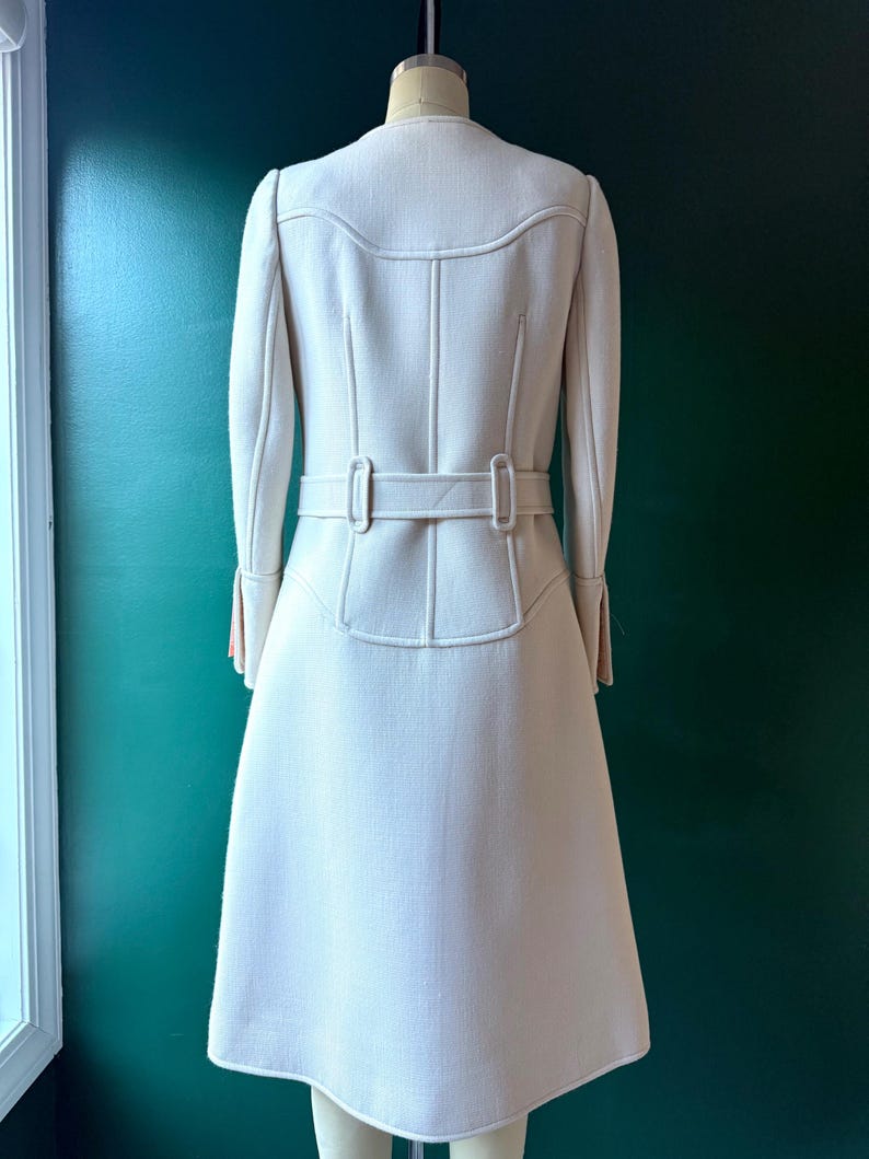 May include: A white coat with a belt and long sleeves. The coat has a fitted waist and a flared skirt.