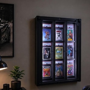 May include: A black wall-mounted display case with integrated LED lighting, featuring three vertical columns that hold nine rectangular trading cards in protective plastic slabs. The case has a dark frame and illuminates the collection of cards.