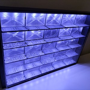 May include: A black, angled display case with multiple clear, rectangular compartments. The compartments are illuminated with a blue light, creating a futuristic aesthetic. The case is likely designed for showcasing small items or collectibles.