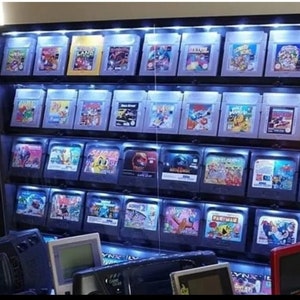 May include: A wall shelf filled with rows of video game cartridges. The cartridges are organized by color and have a variety of titles. The shelf is lit with white lights, highlighting the colorful cartridges.