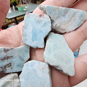 May include: A collection of rough-cut larimar stones, showcasing a mix of pale blue and white hues with natural patterns. The stones vary in size and shape, with some displaying darker gray inclusions. The image is taken in natural light.