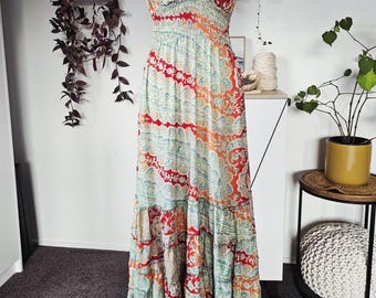Könnte beinhalten: Ein langes, fließendes Kleid mit einem farbenfrohen Paisley-Muster in Rot, Orange und Türkis. Das Kleid hat einen gerüschten Ausschnitt und einen Stufenrock. Der Stoff wirkt leicht, geeignet für warmes Wetter. Das Kleid wird auf einer Büste präsentiert.