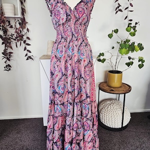 May include: A long, flowing dress with a vibrant paisley pattern in shades of pink, purple, and blue. The dress features a V-neckline, ruffled tiers, and a fitted bodice. The dress is displayed on a mannequin.