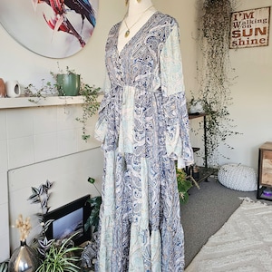 May include: A long, flowing blue and white paisley print maxi dress with a V-neckline, long sleeves, and a tiered skirt. The dress is cinched at the waist with a tie.