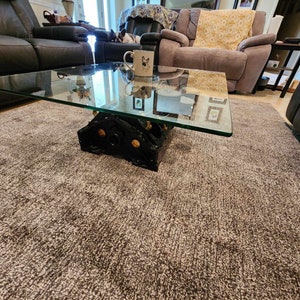 May include: A glass-top coffee table with a unique industrial base made from repurposed engine parts. The table sits on a gray shag rug in a living room setting.