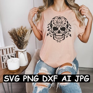 Może przedstawiać: R&oacute;żowy t-shirt z czarno-białym wzorem czaszki z kwiatami. Tekst "SVG PNG DXF AI JPG" jest nadrukowany na czarnym banerze pod obrazkiem.
