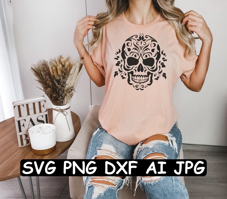 Może przedstawiać: R&oacute;żowy t-shirt z czarno-białym wzorem czaszki z kwiatami. Tekst "SVG PNG DXF AI JPG" jest nadrukowany na czarnym banerze pod obrazkiem.