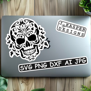 Może przedstawiać: Czarno-biała czaszka z kwiatowymi wzorami, na srebrnym laptopie z białą naklejką z napisem "SVG PNG DXF AI JPG" i inną białą naklejką z napisem "J. Kasper Designs".