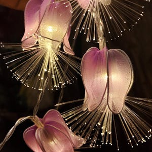 May include: A string of decorative lights featuring multiple light purple, bell-shaped flower petals. Each flower is illuminated from within and surrounded by thin, radiating fibre optic filaments that emit a bright white glow at their tips.