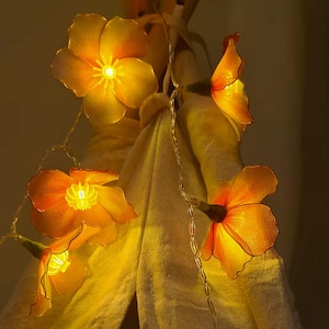 Puede incluir: Una cadena de luces decorativas con forma de flores de color naranja-amarillo. Las luces están ensartadas en un fino cable plateado. Las flores tienen una cualidad translúcida, con una luz brillante que emana de sus centros. El fondo es de un color suave y neutro.