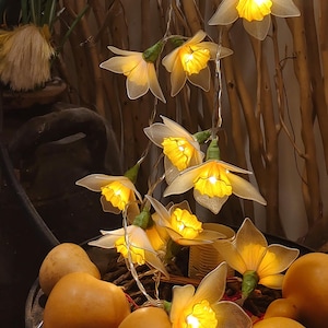 Puede incluir: Una cadena de luces decorativas con forma de flor, cada una con un cálido brillo amarillo. Las flores tienen pétalos translúcidos y tallos verdes. Las luces cuelgan frente a un fondo de ramas de madera y calabazas.