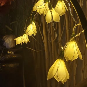 May include: A string of LED fairy lights featuring translucent, pale yellow flower-shaped shades. Each flower has delicate, petal-like structures with a fine, grid-like texture. The lights emit a warm, soft glow, connected by a thin, flexible wire.
