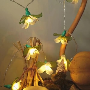 Könnte beinhalten: Eine Lichterkette mit weißen, blumenförmigen Glühbirnen und grünen Blättern. Die Lichter sind um eine kleine Anordnung aus getrockneten Zweigen, einem großen Stein und anderen natürlichen Elementen gespannt.