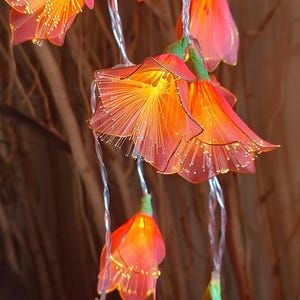 May include: A string of decorative flower-shaped lights featuring translucent coral-pink petals with gold-toned edges. Each flower contains a bright yellow LED centre with thin, radiating filaments that glow, connected by a thin, flexible silver wire.
