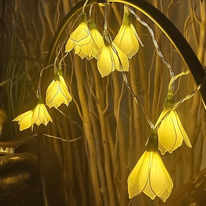 Könnte beinhalten: Eine LED-Lichterkette mit durchscheinenden, blassgelben blütenförmigen Abdeckungen. Jede Blüte hat einen grünen Sockel und ist mit einem dünnen, flexiblen Draht verbunden. Die Lichter strahlen ein warmes, sanftes Licht aus, das die zarte Textur betont.