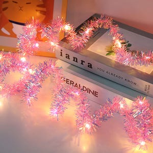 May include: Pink tinsel garland with small, warm white lights. The garland is draped over a stack of books with the titles "ianra" and "Geraldine". The lights are evenly spaced along the garland, creating a festive look.