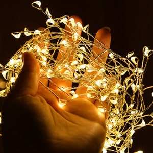 Pu&ograve; includere: Una mano che tiene una stringa di luci bianche calde. Le luci sono avvolte attorno alla mano e brillano intensamente.
