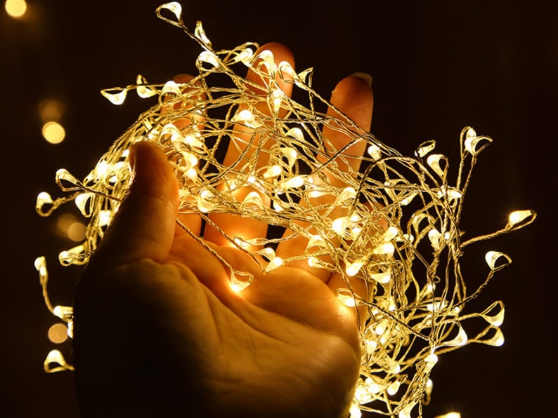 Pu&ograve; includere: Una mano che tiene una stringa di luci bianche calde. Le luci sono avvolte attorno alla mano e brillano intensamente.