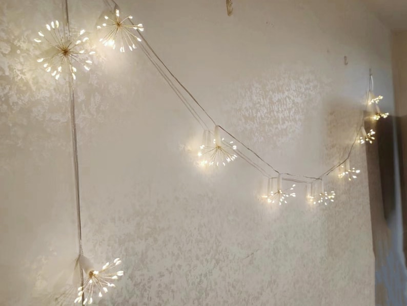 K&ouml;nnte beinhalten: Eine Lichterkette mit wei&szlig;en Feenlichtern im Sternburst-Design. Die Lichter sind auf einem durchsichtigen Draht aufgef&auml;delt und h&auml;ngen an einer wei&szlig;en Wand.