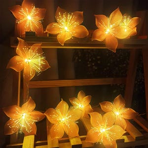 Può includere: Ghirlanda di luci con un designo a forma di fiore. Ogni luce è un piccolo fiore giallo con un centro in fibra ottica bianca. Le luci sono infilate su un filo trasparente e sono appese a uno scaffale di legno.