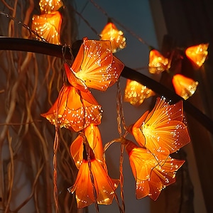 Può includere: Una stringa di luci a forma di fiore arancione caldo. Ogni fiore ha un design traslucido a forma di petalo con piccole luci bianche che emanano dal centro. Le luci sono infilate su un filo sottile e scuro, creando un bagliore morbido e ambientale.