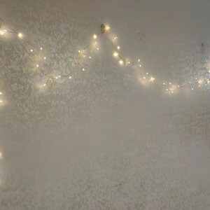 Pu&ograve; includere: Una ghirlanda di luci bianche a forma di arco, appesa a un muro bianco. Le luci creano un'atmosfera festosa e fantasiosa.