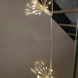 K&ouml;nnte beinhalten: Zwei wei&szlig;e Lichterketten mit einem B&uuml;ndel kleiner wei&szlig;er Lichter am Ende jeder Kette. Die Lichter sind in Form eines Feuerwerks oder eines L&ouml;wenzahns geformt.