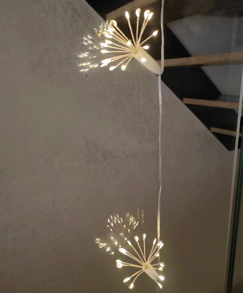 K&ouml;nnte beinhalten: Zwei wei&szlig;e Lichterketten mit einem B&uuml;ndel kleiner wei&szlig;er Lichter am Ende jeder Kette. Die Lichter sind in Form eines Feuerwerks oder eines L&ouml;wenzahns geformt.