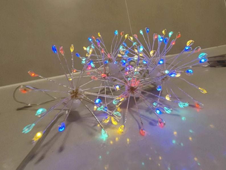 K&ouml;nnte beinhalten: Ein Satz mit drei mehrfarbigen LED-Lichterketten in Form einer Feuerwerksfont&auml;ne. Die Lichter sind auf einem klaren Draht aufgef&auml;delt und haben verschiedene Farben, darunter Rot, Blau, Gr&uuml;n, Gelb und Wei&szlig;.